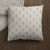 A square throw pillow with a light beige linen-like fabric and a repeating small black hand block floral pattern, resting on a dark brown wooden floor next to a plaid blanket.