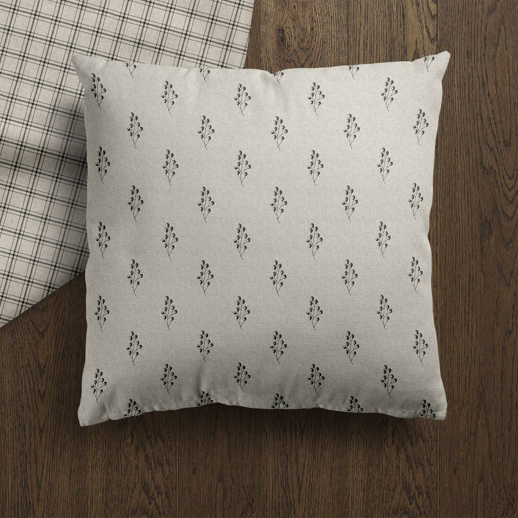 A square throw pillow with a light beige linen-like fabric and a repeating small black hand block floral pattern, resting on a dark brown wooden floor next to a plaid blanket.