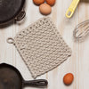 Textured pot holder on a wooden surface with eggs and a whisk.