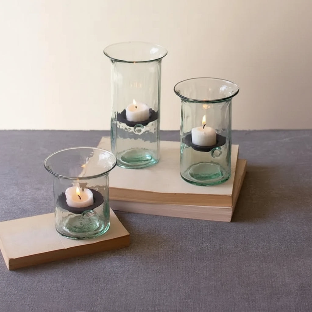 Three glass candle holders with candles on a gray surface, with a plant in the background.