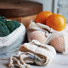 Mesh produce bags with oranges and broccoli on a wooden surface with a plant in the background.