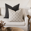 Two decorative throw pillows on a light beige sofa in a styled living room. The front pillow is cream with a small repeating dark gray botanical pattern, and the back pillow is dark gray with a subtle grid pattern.