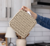 A hand holding a square, chunky-knit olive colored pot holder in a kitchen setting with white subway tiles and light wood cabinets in the background.