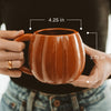 Brown ceramic mug held by hands with measurements indicated, blurred background
