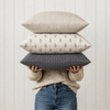 Three decorative throw pillows in different patterns (checkered, patterned, and plaid) are stacked on a person's shoulders who is wearing a cream knit sweater and blue jeans, set against a white paneled wall.