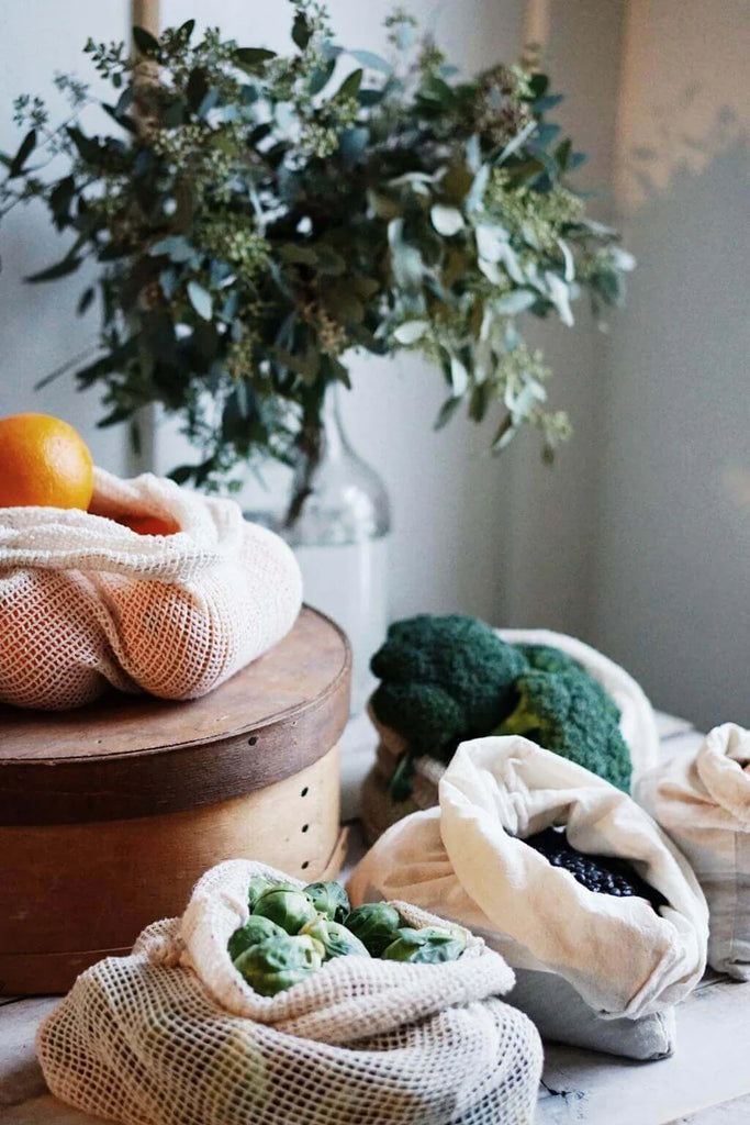 Reusable mesh produce bags with fruits and vegetables on a wooden surface with a plant in the background.