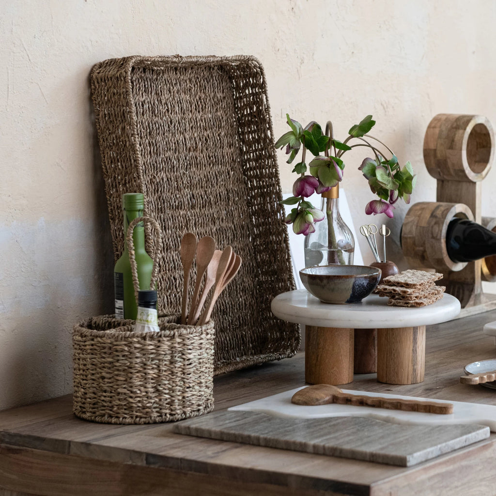 Wooden table setting with utensil caddy, bottles, and decorative items against a neutral wall.