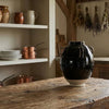 A large glossy black ceramic vase with two handles and a white foot ring sits on a rustic wooden kitchen island. The background shows a warm, traditional kitchen with white shelves holding dishes, copper pots, and dried herbs hanging near a window.