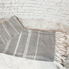 A gray and white striped woven turkish bath towel with fringed edges, draped on a light stone surface against a white brick wall.