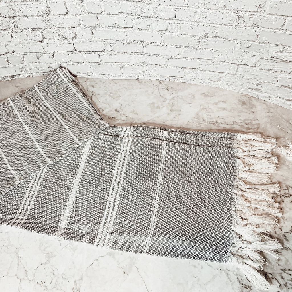 A gray and white striped woven turkish bath towel with fringed edges, draped on a light stone surface against a white brick wall.
