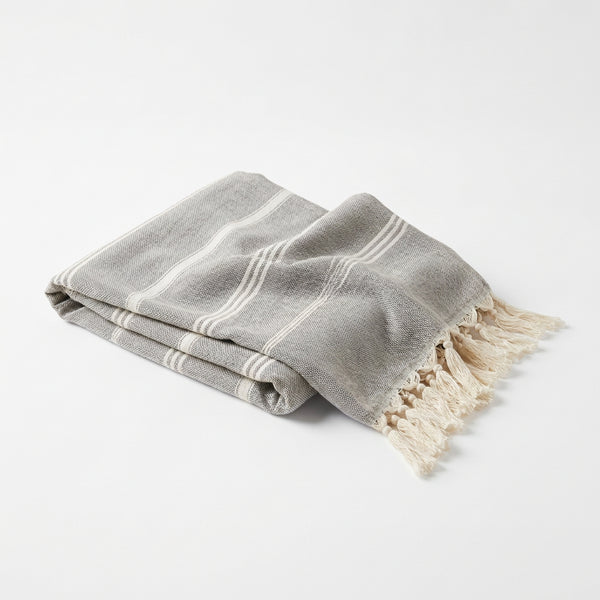 A gray and white striped woven turkish bath towel with fringed edges, draped on a light stone surface against a white brick wall.