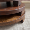 Three round wooden pedestals stacked together on a light beige textured fabric surface, with a softly blurred indoor background.