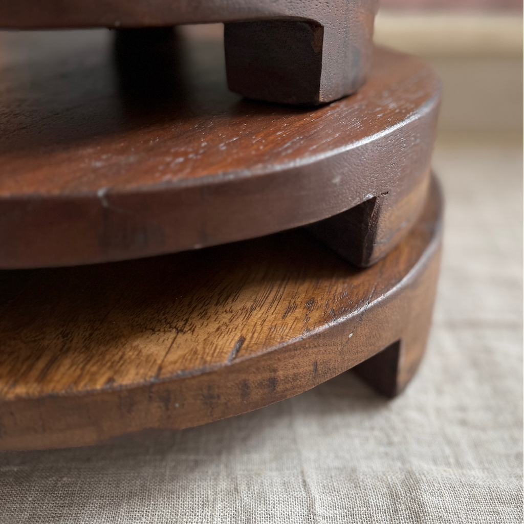 Three round wooden pedestals stacked together on a light beige textured fabric surface, with a softly blurred indoor background.
