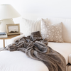 A plush, soft gray throw blanket is casually draped over a white sofa in a bright, modern living room setting, styled with neutral patterned and striped pillows, a side table with a lamp, and a picture frame.