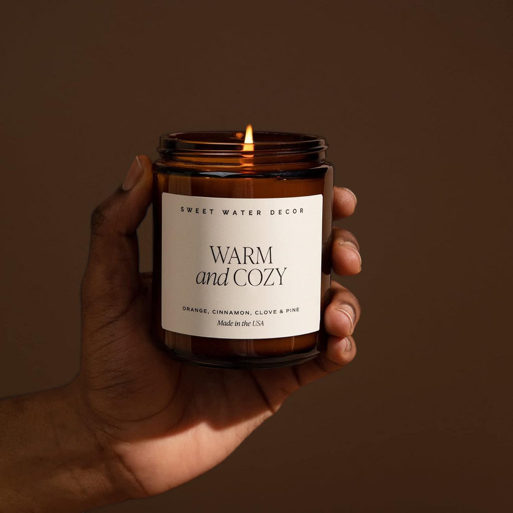 Hand holding a lit candle labeled 'Warm and Cozy' against a brown background