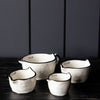 Four white ceramic measuring cups with black rims and handles, arranged on a dark wooden surface against a dark gray paneled background.