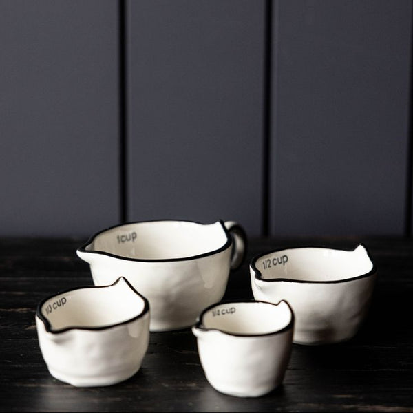 Four white ceramic measuring cups with black rims and handles, arranged on a dark wooden surface against a dark gray paneled background.
