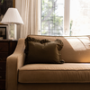 A beige upholstered sofa in a warmly lit living room, styled with a dark olive green ruffled throw pillow. A white pleated table lamp sits on a dark wooden side table to the left, and a bright window with cream curtains is in the background.