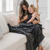 Woman and child hugging under a dark gray blanket on a couch in a bright room.