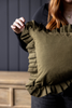 An olive green fabric throw pillow with ruffled trim, held by a person in a black top against a dark paneled wall and wooden furniture background.