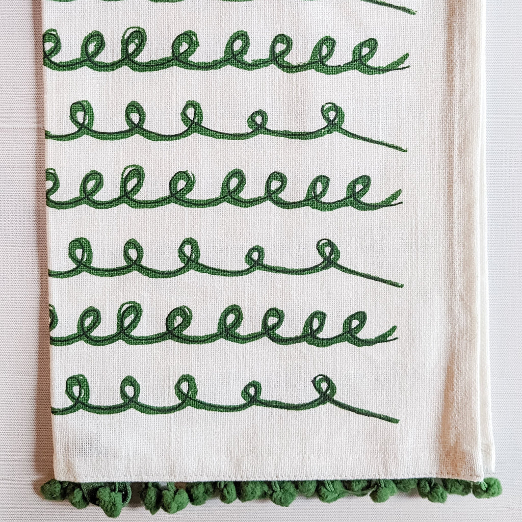 A white cotton tea towel with green swirls and pom poms decoration, displayed on a white background.