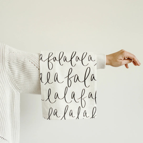A person is holding up a cream-colored kitchen towel with black hand-lettered text 'Fa La La La La'.