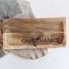 A mango wood serving board with 'Give Thanks' carved on the side, placed on a textured surface.
