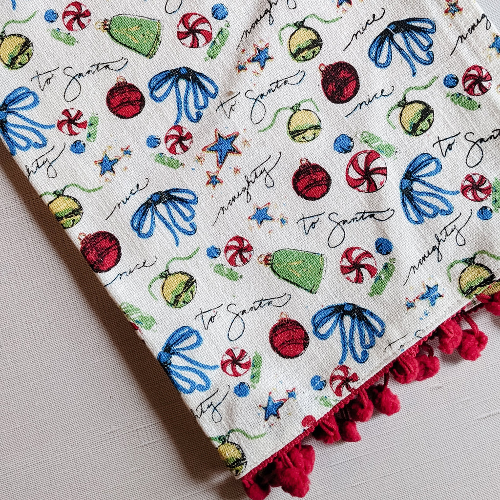 A white Christmas-themed tea towel with multicolored Christmas decoration prints, pom-pom edging, and a red loop for hanging.