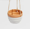 A small white hanging planter made of clay, with a terracotta-colored bottom and a white upper part, suspended by a jute twine.