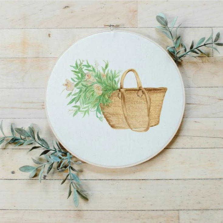 A watercolor hoop with a floral market bag design, featuring a beige background and green and yellow floral patterns, placed on a wooden surface with a few green leaves around it.