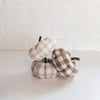 Three mini pumpkin-shaped fabric pillows in checkered patterns of beige, white, and brown colors, arranged against a white brick wall background.