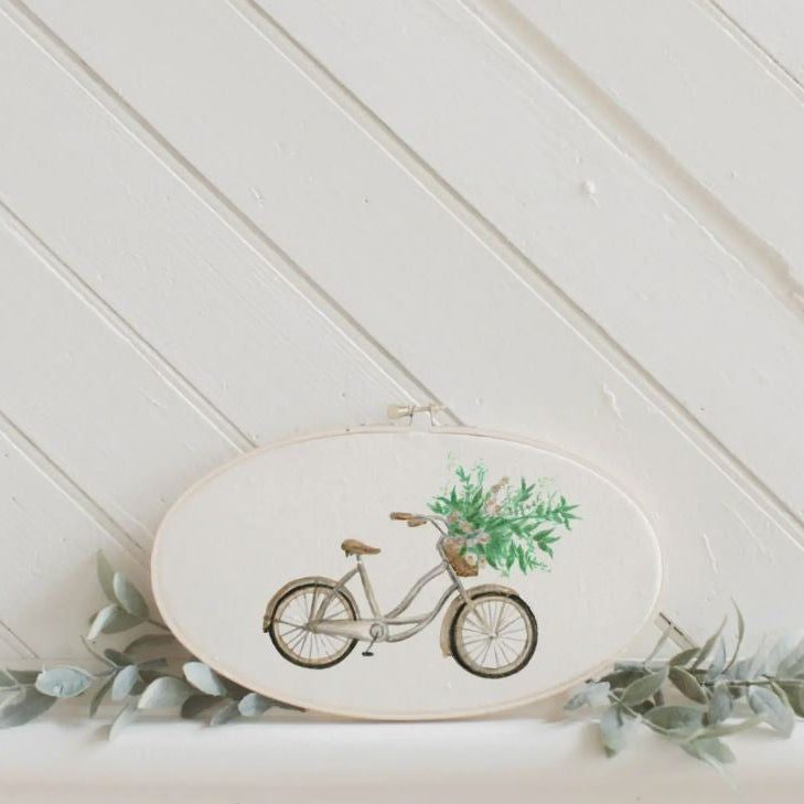 A watercolor hoop with a floral bike design, placed against a white wooden background with a small greenery arrangement beside it.